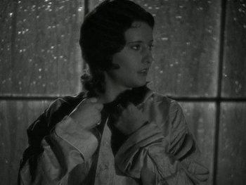 Movie still from “Ladies of Leisure” (1930), directed by Frank Capra – A young woman in a dress adjusting her neck tie in front of a window; Medium shot, Low angle