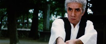 Movie still from “Lady Snowblood” (1973), directed by Toshiya Fujita – An older man in a white and black shirt is holding his hands out; Close Up shot, Low angle