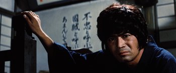 Movie still from “Lady Snowblood” (1973), directed by Toshiya Fujita – A man in a hat is sitting in front of a wall with writing on it; Close Up shot, Low angle