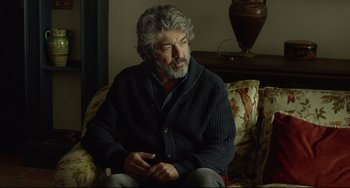 Movie still from “Everybody Knows” (2018), directed by Asghar Farhadi – An older man sitting on a couch in a living room; Medium shot, Over the shoulder angle