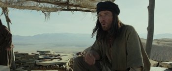 Movie still from “Last Days in the Desert” (2015), directed by Rodrigo García – A man with long hair sitting on the ground in the desert; Medium shot, Over the shoulder angle