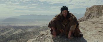 Movie still from “Last Days in the Desert” (2015), directed by Rodrigo García – A man sitting on top of a rock on top of a mountain; Medium shot, Low angle