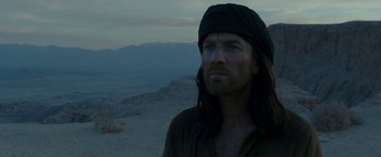 Movie still from “Last Days in the Desert” (2015), directed by Rodrigo García – A man with long black hair wearing a black hat; Close Up shot, Over the shoulder angle