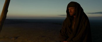 Movie still from “Last Days in the Desert” (2015), directed by Rodrigo García – A man in a hooded cloak sitting on top of a hill; Medium shot, Low angle