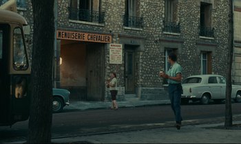 Movie still from “Happiness” (1965), directed by Agnès Varda – A woman walking down the street while a man is walking down the sidewalk; Wide shot, Over the shoulder angle