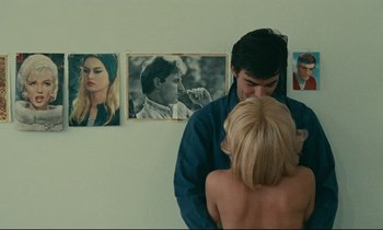 Movie still from “Happiness” (1965), directed by Agnès Varda – A man and a woman standing next to each other in front of pictures; Medium shot, Over the shoulder angle