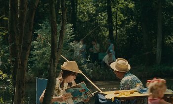 Movie still from “Happiness” (1965), directed by Agnès Varda – A woman reading a magazine in front of a group of people in the woods; Wide shot, Over the shoulder angle