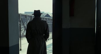 Movie still from “Le Samouraï” (1967), directed by Jean-Pierre Melville – A man in a trench coat and a hat looking out a window; Wide shot, Over the shoulder angle