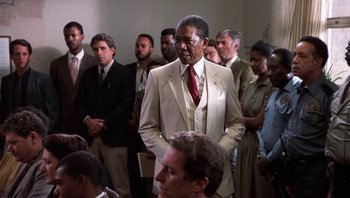 Movie still from “Lean on Me” (1989), directed by John G. Avildsen – A group of men in suits and ties standing in a room; Medium shot, Over the shoulder angle