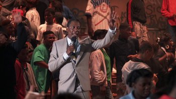 Movie still from “Lean on Me” (1989), directed by John G. Avildsen – A man is holding a microphone and waving at a crowd; Medium shot, Low angle