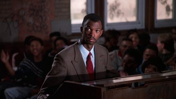 Movie still from “Lean on Me” (1989), directed by John G. Avildsen – A man in a brown suit and a red neck tie is speaking; Medium shot, Low angle