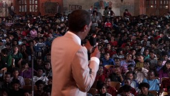 Movie still from “Lean on Me” (1989), directed by John G. Avildsen – A man in a white suit is holding a microphone in front of a crowd; Medium shot, High angle