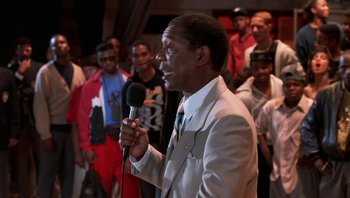 Movie still from “Lean on Me” (1989), directed by John G. Avildsen – A man holding a microphone in front of a group of people; Medium shot, Over the shoulder angle