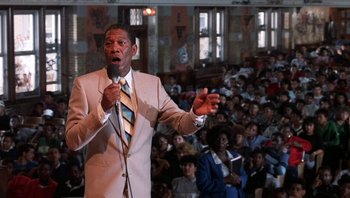 Movie still from “Lean on Me” (1989), directed by John G. Avildsen – A man in a tan suit is holding a microphone; Medium shot, Low angle