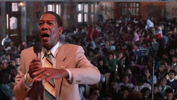 Movie still from “Lean on Me” (1989), directed by John G. Avildsen – A man in a white suit is holding a microphone; Medium shot, Low angle