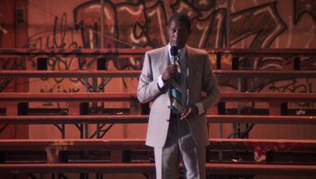 Movie still from “Lean on Me” (1989), directed by John G. Avildsen – A man wearing a suit and tie holding a microphone; Medium shot, Low angle