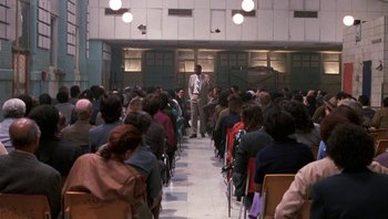 Movie still from “Lean on Me” (1989), directed by John G. Avildsen – A man standing in front of a crowd of people in a room; Wide shot, High angle