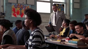 Movie still from “Lean on Me” (1989), directed by John G. Avildsen – A man in a suit and tie standing in front of a group of people; Medium shot, Over the shoulder angle