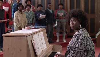Movie still from “Lean on Me” (1989), directed by John G. Avildsen – A woman sitting at a piano in front of a group of people; Medium shot, High angle