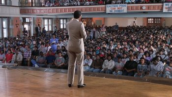 Movie still from “Lean on Me” (1989), directed by John G. Avildsen – A crowd of people sitting and standing in front of a man on a stage; Wide shot, High angle