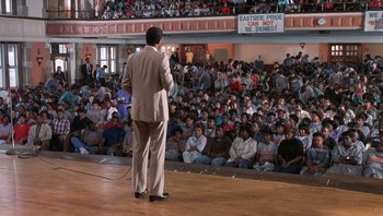 Movie still from “Lean on Me” (1989), directed by John G. Avildsen – A man standing on a stage in front of an audience; Wide shot, High angle