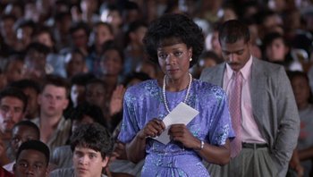Movie still from “Lean on Me” (1989), directed by John G. Avildsen – A woman in a purple dress holding a piece of paper in front of a crowd of people; Medium shot, High angle
