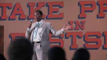 Movie still from “Lean on Me” (1989), directed by John G. Avildsen – A man in a gray suit is holding a microphone; Medium shot, Low angle