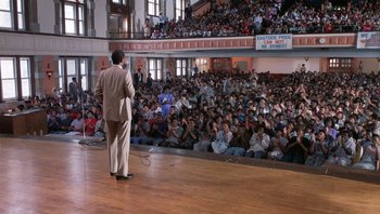 Movie still from “Lean on Me” (1989), directed by John G. Avildsen – A man is speaking on a stage in front of a crowd; Wide shot, High angle