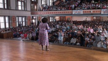 Movie still from “Lean on Me” (1989), directed by John G. Avildsen – A woman in a colorful dress standing in front of an audience; Extreme Wide shot, High angle