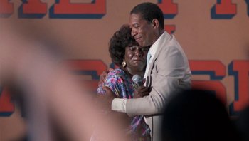 Movie still from “Lean on Me” (1989), directed by John G. Avildsen – An older man hugging a woman while holding a microphone; Close Up shot, Over the shoulder angle