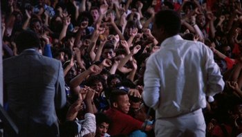 Movie still from “Lean on Me” (1989), directed by John G. Avildsen – A group of people sitting in front of an audience with their hands up; Wide shot, High angle