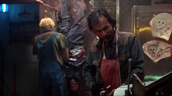 Movie still from “Leatherface: Texas Chainsaw Massacre III” (1990), directed by Jeff Burr – A man wearing an apron is working in a workshop; Medium shot, Over the shoulder angle