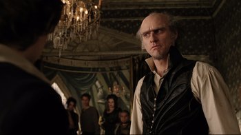 Movie still from “A Series of Unfortunate Events” (2004), directed by Brad Silberling – A man in a black and white shirt and a group of people in a room; Medium shot, Over the shoulder angle