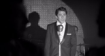 Movie still from “Lenny” (1974), directed by Bob Fosse – A man in a tuxedo standing next to a microphone in front of an audience; Medium shot, Over the shoulder angle