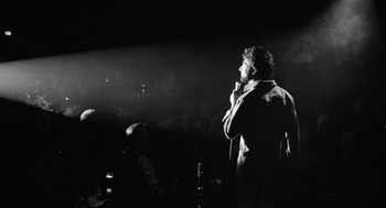 Movie still from “Lenny” (1974), directed by Bob Fosse – A man standing on a stage holding a microphone; Medium shot, Low angle