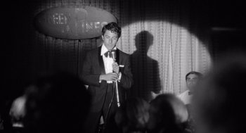 Movie still from “Lenny” (1974), directed by Bob Fosse – A black and white photo of a man in a suit holding a microphone; Medium shot, Over the shoulder angle