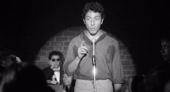 Movie still from “Lenny” (1974), directed by Bob Fosse – A man standing on a stage holding a microphone; Medium shot, Low angle