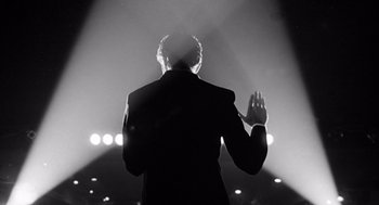 Movie still from “Lenny” (1974), directed by Bob Fosse – A man standing in front of a stage with his hand raised; Medium shot, Low angle