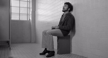 Movie still from “Lenny” (1974), directed by Bob Fosse – A man sitting on top of a piece of luggage in a bathroom; Medium shot, Low angle