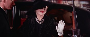 Movie still from “Les Girls” (1957), directed by George Cukor – A woman in a black dress and a black hat waves to onlookers; Close Up shot, Low angle