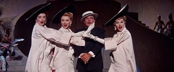 Movie still from “Les Girls” (1957), directed by George Cukor – A man in a black suit and two women in white hats; Medium shot, Low angle