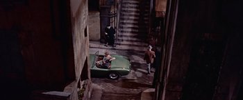 Movie still from “Les Girls” (1957), directed by George Cukor – A man driving a green car down a street; Wide shot, High angle
