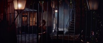 Movie still from “Les Girls” (1957), directed by George Cukor – A man sitting in front of a metal fence; Wide shot, High angle
