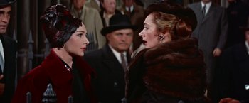 Movie still from “Les Girls” (1957), directed by George Cukor – Two women in fur coats talking to each other in front of a crowd of people; Close Up shot, Over the shoulder angle