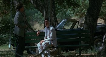 Movie still from “Less Than Zero” (1987), directed by Marek Kanievska – A man sitting on top of a wooden bench next to a tree; Wide shot, Over the shoulder angle