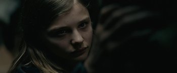 Movie still from “Let Me In” (2010), directed by Matt Reeves – A young girl looking at the camera in a dimly - lit room; Close Up shot, Over the shoulder angle