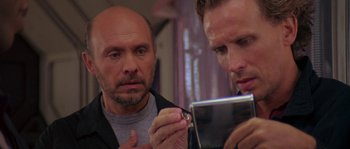 Movie still from “Leviathan” (1989), directed by George P. Cosmatos – A man looking at his reflection in a mirror while another man holds a cell phone; Close Up shot, Over the shoulder angle