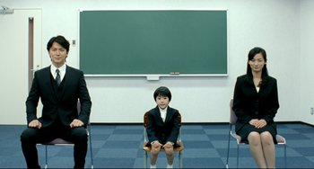 Movie still from “Like Father, Like Son” (2013), directed by Kore-eda Hirokazu – A woman sitting in front of a chalkboard with two men standing next to her; Wide shot, High angle