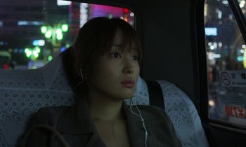 Movie still from “Like Someone in Love” (2012), directed by Abbas Kiarostami – A woman sitting in the back seat of a car wearing headphones; Close Up shot, Over the shoulder angle
