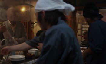 Movie still from “Like Someone in Love” (2012), directed by Abbas Kiarostami – A person wearing a white hat in front of a counter; Medium shot, Over the shoulder angle